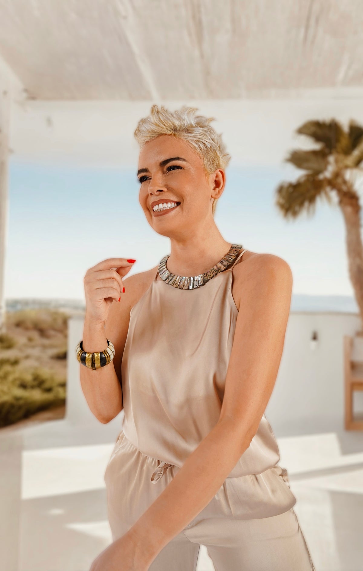 Queen Paua sea shell necklace on a smiling model in beige satin, white terrace with a palm tree