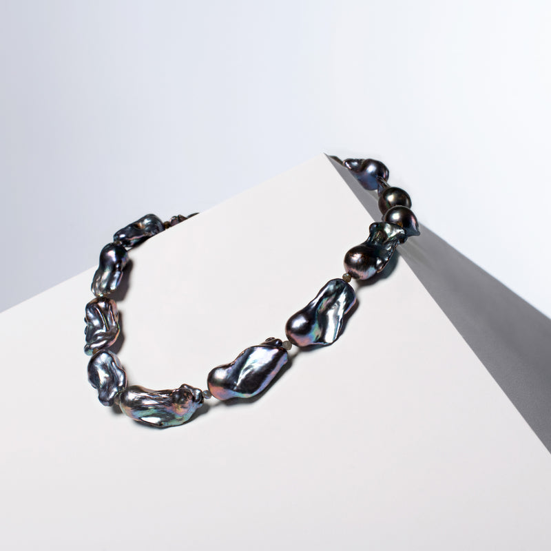 Cosmos black baroque pearl necklace resting on a white angled block, HoS Lock out of view