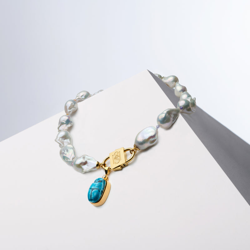 Scarab charm hanging from the HoS Lock of a baroque pearl necklace on a white block